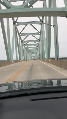 The video shows a first-person view from inside a vehicle driving across a bridge. The bridge has a green, truss-style structure with a clear roadway. There are yellow lines marking the lanes. In the distance, a few other vehicles are visible on the bridge. The sky is overcast and a bit foggy, obscuring the view of the surroundings beyond the bridge. The dashboard of the vehicle is visible at the bottom of the frame.