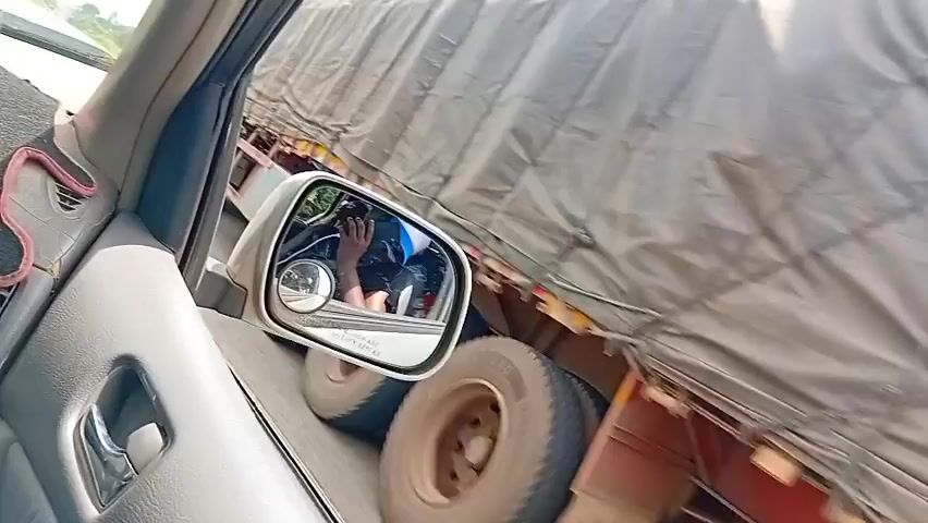 The video begins from inside a vehicle, showing a side mirror reflecting the driver and the interior of the car. The driver is wearing a blue shirt. Outside the vehicle, a large truck covered with a gray tarp is visible. The truck appears to be a flatbed with large tires. The camera then pans to show the road and surrounding environment, which consists of trees and a cloudy sky. As the video progresses, the vehicle passes a red truck. The camera continues to capture the scenery outside, with glimpses of vegetation and utility poles. The video ends with a view of the vehicle's dashboard.