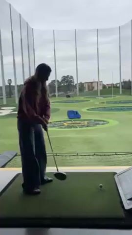 A woman in a maroon sweatshirt and jeans is playing golf at a Topgolf facility. She is standing on a green mat in a hitting bay with a golf club in her hands, preparing to swing at a golf ball on a tee. The facility has netted walls and targets on the field. The sky is overcast.