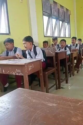 The video shows a group of young boys sitting at desks in a classroom. They are all wearing white shirts and maroon pants, suggesting a school uniform. Most of the boys have backpacks on their chairs or on their backs. The classroom appears to be lit by natural light coming from windows on the far wall. The walls are painted yellow. The desks are made of wood and some have graffiti on them. Some of the boys appear to be looking forward, possibly at a teacher who is not visible in the frame. One boy in the foreground is looking down at his desk, while another boy in the middle ground is looking slightly up and appears to be speaking or singing. The overall impression is of a typical classroom scene during a lesson.