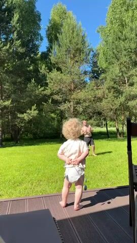 A toddler with curly blonde hair and wearing a diaper stands on a wooden deck with their back to the camera, holding a spoon and fork. They are facing a man with a beard and dark shorts, who is standing on a green lawn with a drink in his hand. The scene is outdoors with many tall green trees under a clear blue sky.