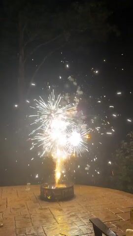 The video shows fireworks being set off at night. The fireworks are bright white and orange, exploding in a shower of sparks. They are being set off from a fire pit on a patio. The patio is made of stone and appears to be wet. There are trees and foliage in the background. The video is shot from a low angle, looking up at the fireworks.