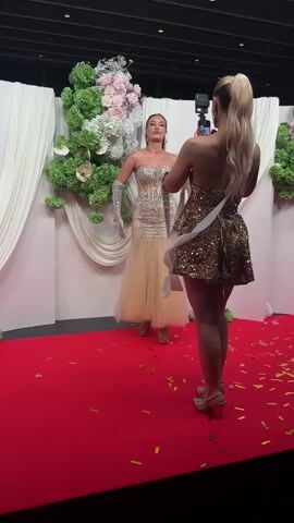 The video captures a red-carpet event where two women, presumably beauty pageant contestants, are interacting. One woman, adorned in a shimmering gold mini-dress and heels, stands on the red carpet with her back to the camera, holding a smartphone in her hands and seemingly taking a photograph. The other woman, positioned directly in front of her, poses for the photo, wearing a stunning strapless gown, long gloves, and matching heels. Both women are wearing sashes, further suggesting their involvement in a pageant.

The environment is decorated with white draped curtains, floral arrangements, and scattered gold confetti on the red carpet, enhancing the glamorous atmosphere. The lighting is bright, illuminating the scene and highlighting the women's attire. The soundscape includes music, adding to the celebratory vibe of the event.