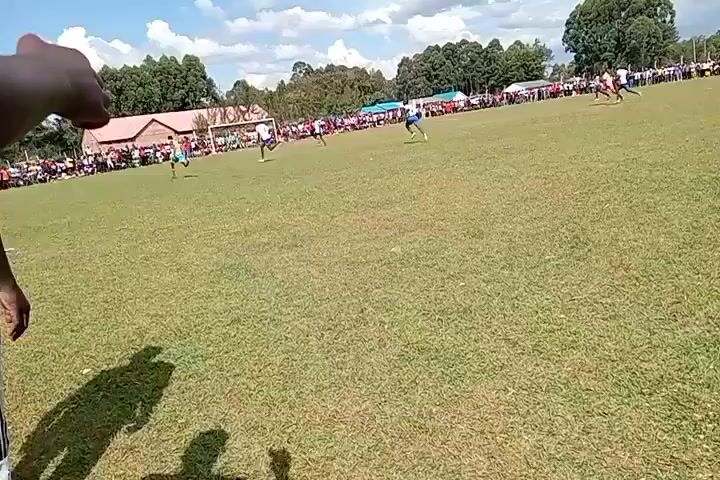 The video shows a soccer game in progress, from the perspective of someone on the sideline. The field is green with players in varying colored uniforms running around. A crowd is visible along the sidelines, watching the game. The sky is blue with scattered white clouds. In the foreground is a person in a white and green shirt, facing the field. The camera is relatively stable, providing a clear view of the game and the surrounding environment.