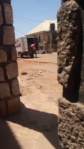 A street scene is captured through a stone archway. The focus is on a white truck parked on the road, where two men are visible near the back of the truck. One man is wearing a red shirt, and the other is partially obscured. The truck has writing on its side. A building is visible in the background, along with a water tank on the roof. The road appears to be unpaved. The sky is blue. A person can be seen sitting in the foreground. The scene seems to be taken during the daytime.