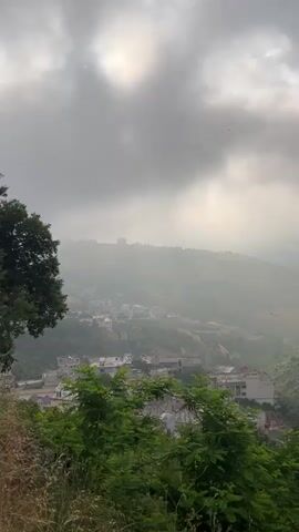 A landscape video shows a view of a town nestled in a valley, surrounded by hills and trees. The sky is overcast with dramatic clouds, and the sun is attempting to break through, casting a hazy light over the scene. The town consists of buildings with white and light-colored facades, some with red roofs, clustered together along the slopes. In the foreground, lush green foliage, including bushes and tall grass, partially obscures the view. The overall impression is one of a slightly misty or foggy morning in a rural or semi-rural mountainous area. There are no people or animals visible in the frame. The video is shot from an elevated vantage point, looking down into the valley.
