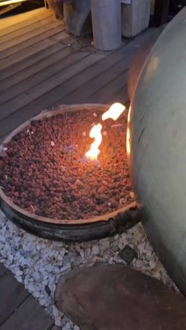 The video shows a small fire pit and a koi pond with orange koi fish. The fire pit is round and contains red rocks, with a small flame burning in the center. A large, rounded ceramic pot sits next to the fire pit. The koi pond is surrounded by lush greenery, including large-leafed plants and grasses. There are blue and yellow lights illuminating the pond. The camera pans from the fire pit to the koi pond.