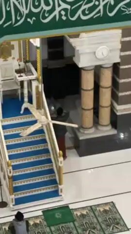The video shows the interior of a mosque. An individual wearing a gray jacket and what appears to be a sarong is standing and facing the entrance to an elevated platform or minbar. The minbar is adorned with blue and gold detailing and has a staircase leading up to it. A ceiling fan hangs above the staircase. In the foreground, a person in a striped shirt is sitting on a green prayer rug on the floor. The floor is white with black accents. Above the entrance, there is green calligraphy. There is a clock above the entrance. The video seems to have been taken from an elevated position, likely a balcony or upper level of the mosque. A man puts his hand on his head.