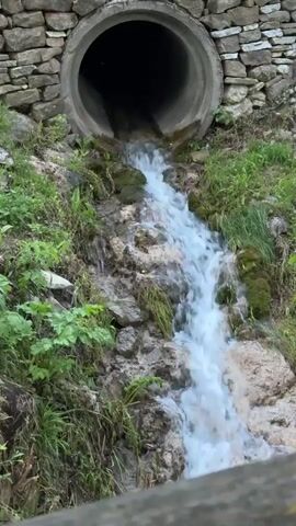 The video shows a small waterfall emanating from a large concrete pipe embedded in a stone wall. The water cascades down a rocky, moss-covered slope, surrounded by green vegetation. The overall scene has a natural, outdoor setting.