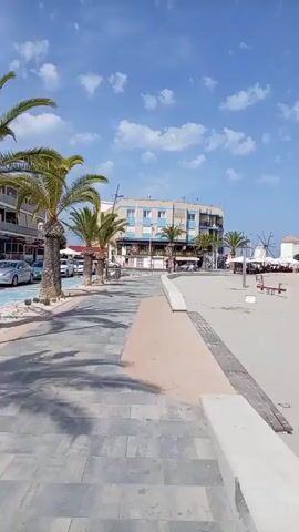 This media file depicts a sunny day at a beach in Villa Nanitos, Spain. The scene is a wide, sandy beach with a clear blue sky dotted with fluffy white clouds. In the foreground, a paved walkway with decorative wooden planks runs along the edge of the beach. Further back, a children's playground with brightly colored slides and swings is visible. Beyond the playground, the sandy beach stretches out, dotted with beach umbrellas and a few scattered people enjoying the day. In the distance, a row of buildings and palm trees line the beachfront. Two traditional windmills, a characteristic feature of the location, stand prominently against the sky. The overall atmosphere appears peaceful and leisurely, suggesting a typical summer day at the seaside. The presence of beachgoers, playground equipment, and umbrellas indicates recreational activity. The bright sunlight and clear sky suggest it is daytime, likely late morning or early afternoon. No visible text is present in the media file.