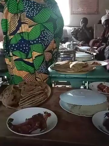 The video shows a room where food is being prepared and served. In the foreground, a table is loaded with plates of food including what appears to be bread, meat, and stews. A woman in a colorful patterned dress stands beside the table and appears to be serving from a pot. In the background, two or three other women are sitting around a table, possibly eating or preparing food. The room seems to be modestly furnished, with a window providing natural light. There's a sign on the wall. The atmosphere appears to be communal, suggestive of a gathering or meal preparation for a group.