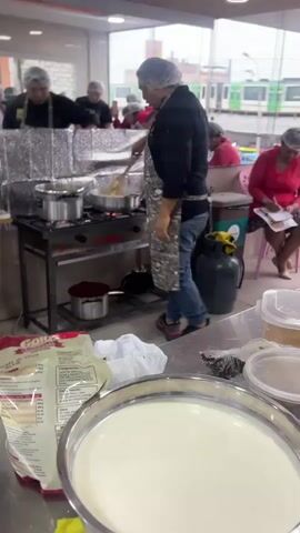A cooking class is in session. A male instructor, wearing a black chef's coat, a patterned apron, and a hairnet, stands over a four-burner gas stove, stirring a pot. He is facing a group of students, all wearing hairnets, some of whom are watching him. To the right, a woman in a red shirt is seated, looking at notes. In the foreground, there are large metal bowls and bags of ingredients. To the left, a woman in a purple apron is pointing to a recipe card. The room appears to be a commercial kitchen or classroom with a large window in the background.