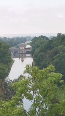 The video pans across a wide, calm river surrounded by dense green trees. In the distance, a stone bridge with architectural features, possibly a dam or aqueduct, spans the river. The sky is overcast, contributing to a soft, natural light. The foreground is partially obscured by the leaves of a tree, adding depth to the scene. The overall impression is one of natural beauty and a historical structure integrated into the landscape.
