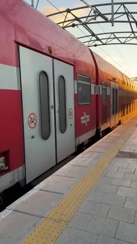 A red and white double-decker train is stopped at a platform. The train has several windows and doors, and a logo with two curved lines is visible on the side. The platform is paved with light-colored tiles and has a yellow tactile paving strip along the edge. Overhead, there are power lines for the train. The sun is setting, casting a warm glow on the train and the platform.
