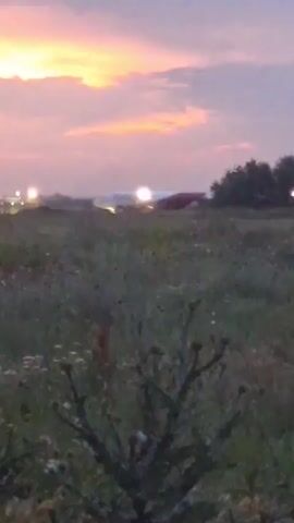 The video shows an airplane with flashing red lights on what appears to be a runway at dusk. The sky is colored with hues of pink, orange, and purple from the sunset. In the foreground, there is a field of green grass and some dark foliage. The airplane is white with red and possibly gold accents on its tail. There are some other lights visible in the distance, possibly from airport infrastructure. The camera is low to the ground, looking up at the airplane. The video is shaky and somewhat blurry, suggesting it was filmed handheld.