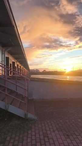 A vertical video captures a sunset over a body of water. In the foreground, a red brick walkway is visible, leading to a grey concrete barrier. To the left, a covered walkway with a red railing and stairs extends into the frame. Beyond the barrier, the water reflects the warm hues of the setting sun. In the background, streetlights line a distant road, and clouds painted with shades of orange, pink, and yellow fill the sky. The sun, a bright orb, is low on the horizon, casting a warm glow across the scene. Buildings and trees are silhouetted against the colorful sky.