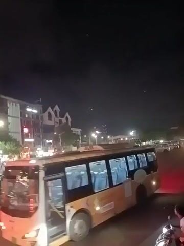 The video shows a static shot of an urban street scene at night. The primary focus is a yellow and brown city bus with "JKSuper Strong" advertisement on its side. The bus is stationary, with its interior seats visible through the windows. In the background, there are several buildings, including a larger one with a gabled roof design, illuminated by streetlights. The sky is dark, and some streetlights are visible. There are also a few motorcycles parked nearby. The camera is stationary, and there are no significant interactions or movements within the frame.