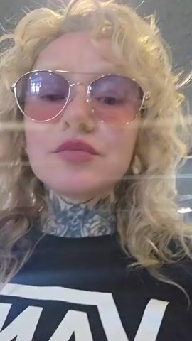 A woman with blonde, curly hair and pink-tinted sunglasses is filming herself. She is wearing a black t-shirt with a white Vans logo. She has tattoos on her neck and arm. Behind her, a man with brown hair and a beard is visible, wearing a black t-shirt with a skull graphic. They appear to be in an indoor public space, possibly an airport or a food court, with displays and seating in the background. The woman is pouting and looking at the camera.