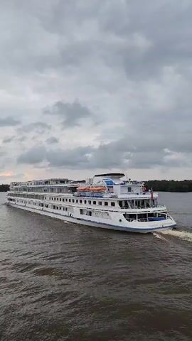 A large white cruise ship sails down a wide river under a cloudy sky. The ship has multiple decks with many windows and balconies. Lifeboats are visible on the upper deck. The water is choppy and brown, with a wake trailing behind the ship. The riverbanks are lined with trees. Another boat, partially visible in the foreground on the left, is following the cruise ship.