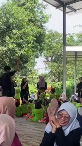 The video shows a group of young children sitting on the ground in rows, wearing green shirts with red pants. They are outside, under a metal structure. In front of them are four adults, three of whom are women wearing hijabs. One woman is wearing glasses and is in the foreground, smiling. The adults appear to be instructing or entertaining the children, with one man holding up a green item and gesturing with his hands. Another adult is taking photos. A large tree is visible in the background.