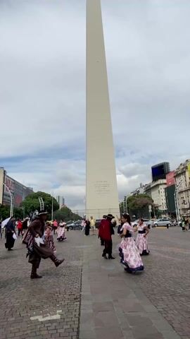 The video shows a group of people dancing in a public square, with the Obelisco de Buenos Aires in the background. The dancers, both men and women, are wearing traditional clothing. The women are wearing long dresses with floral patterns, and the men are wearing dark trousers and hats. The square is paved with cobblestones and there are buildings and traffic visible in the background. The sky is overcast with white clouds. The people are dancing in a large circle to traditional music.