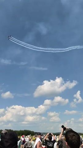 A video shows a blue sky with scattered clouds. In the distance, three planes fly in formation, leaving white smoke trails. The trails form a curving arc across the sky. In the foreground, the heads and shoulders of a crowd of people are visible, looking up at the sky. The people appear to be gathered outdoors, possibly at an airshow or event.
