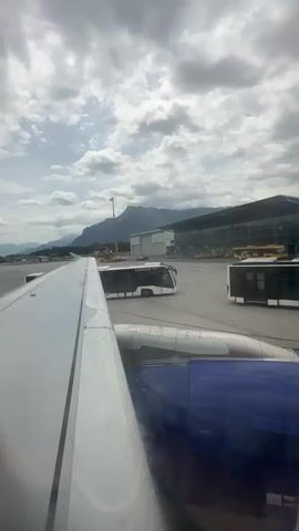 The video shows an aerial view of an airport tarmac from the perspective of an airplane window. In the foreground, the white wing of the airplane is visible, along with part of its engine and a blue section of the fuselage. The tarmac is paved and gray, with several white buses parked at various locations. One bus, labeled "Edubus", is more prominent. In the background, a large modern airport terminal building with a glass facade is visible. Beyond the terminal, mountains rise against a cloudy sky. The overall lighting suggests it is daytime, likely overcast. There are no visible people in motion, except for a very distant figure on the tarmac near a bus. The scene appears static, likely indicating the plane is stationary on the ground.