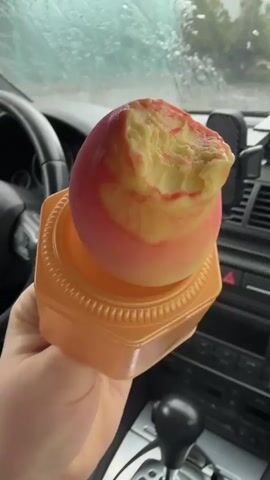 A person is holding a partially eaten ice cream cone inside a car. The ice cream is swirled with pink/red and yellow/cream colors, and sits in an orange, square-based cone. The car's interior is visible, including a black steering wheel on the left, a dashboard, and a gear shifter. Raindrops are visible on the car's windshield, indicating it is raining outside. A phone holder is attached to the dashboard. The car appears to be stationary or moving slowly, with an outdoor background vaguely visible through the wet windshield.