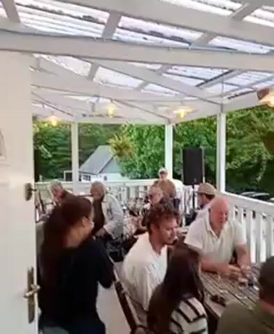 This video captures an outdoor gathering, likely at a cafe or restaurant, on a bright day with a clear sky. The setting is an open-air patio or deck, covered by a white, slatted roof that allows sunlight to filter through. Lush green trees and foliage form a backdrop, with a glimpse of a small house visible among them.

Several people are present, seated at wooden tables or standing. In the foreground, a group is gathered around a table, engaged in conversation. One woman in the center foreground is looking to her left, her expression somewhat obscured. A man to her right, wearing a white polo shirt, is looking towards the table. Another person in the foreground, a woman with dark hair, is wearing a striped shirt.

In the background, a small band appears to be performing. Two individuals are visible playing guitars, and another person seems to be singing or speaking. Two black speaker cabinets are positioned on either side of the band.

The atmosphere seems relaxed and social, suggesting a pleasant afternoon outing. The lighting indicates it is daytime, likely late afternoon or early evening, judging by the warm quality of the light. There is no visible text in the video.