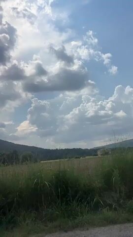 A vertical, first-person perspective video captures a scenic view from inside a vehicle traveling along a road. The sky is partially obscured by dramatic, billowing white and grey clouds, suggesting a dynamic weather system. Sunlight breaks through in some areas, creating bright patches against the blue sky. In the middle ground, a wide, dry, golden field stretches across the landscape, bordered by a line of trees. Beyond the field, rolling hills covered in dense green foliage rise up, creating a layered effect. A small, tan-colored building is visible on the right side of the frame, nestled amongst the trees on the hillside. The foreground of the video is dominated by the interior of the car, including the edge of the dashboard and a silver side-view mirror, which reflects a portion of the sky and the passing scenery. The road itself is visible in the lower left corner, a dark grey asphalt surface. The overall impression is one of a rural, natural landscape experienced during a car journey.