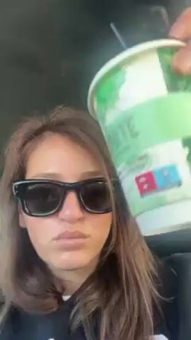 A woman wearing black sunglasses and a black sweatshirt is sitting in a car. She holds up a green and white cup with a black straw. She says "Freddo espresso" in what seems like Greek and takes a sip.