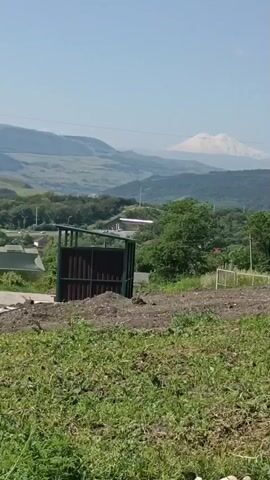 The video shows a landscape with mountains in the background. In the foreground, there is a patch of grass, and a dirt area with a dark metal gate. A road with a white line and a green structure are visible on the left side of the frame. Trees and hills are present in the middle ground. The sky is clear and blue. The video appears to be taken on a sunny day.