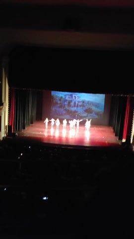 A staged play is in progress, viewed from an elevated angle. The backdrop is a projected image of a pastel-colored castle. In the center of the stage, a group of performers, silhouetted and glowing with white light, are arranged in a diagonal line. They appear to be dressed in light-colored, possibly white, attire. The stage is framed by dark curtains on either side. Red fabric drapes are visible above the stage and along the side. The lighting is dim, with spotlights focused on the performers and the projected backdrop. The audience, or part of it, is visible in the lower right corner, with small, glowing lights indicating their presence.