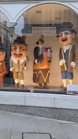 The video shows a storefront window display with five costumed figures standing on platforms. The figures are stylized, with oversized heads and detailed clothing. The text above the display reads "OS CABEZUDOS DE LAXEIRO A Familia Romea." The window reflects the buildings across the street. The camera moves slightly to the side, revealing more of the display. The sidewalk is visible in the foreground.