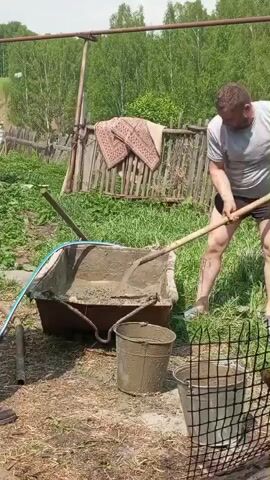 The video shows a man mixing concrete in an outdoor setting. The man, wearing a gray shirt and black shorts, uses a shovel to mix the concrete in a large, rusty trough. A blue hose is connected to the trough, presumably to add water to the mix. There are two buckets nearby, both coated in cement residue. The ground around the trough is a mix of dirt and grass. In the background, there is a rickety wooden fence with patterned rugs hanging on it, and beyond that, a line of trees. The day appears bright and sunny.