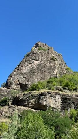 The video shows a conical, rocky mountain peak against a clear blue sky. Green trees and foliage are visible on the lower slopes of the mountain. The camera appears to be moving, as indicated by the slight blurring and shift in perspective throughout the short clip. There are no people or man-made objects clearly visible.
