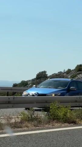 A car and a van drive past a rocky, vegetated hillside alongside a highway under a clear blue sky.