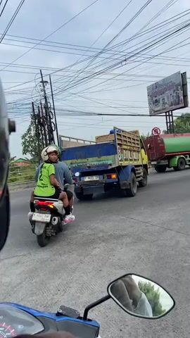 The video shows a street scene from the perspective of a motorcycle. Several vehicles are visible, including a blue dump truck, a green and red water tank truck, and a black car. There are also several motorcycles and pedestrians in the video. The scene appears to be in an urban or suburban area, with buildings and trees lining the street. Traffic signals are present, and a pedestrian crossing is visible on the road. The sky is overcast.
