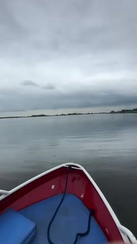 A first-person perspective video shows the bow of a small red and white boat with a blue interior. The boat is on a large body of water with a calm surface reflecting the overcast sky. A dark blue rope is coiled in the bottom of the boat. In the distance, a flat, tree-lined horizon is visible with a few small structures. The sky is filled with heavy, gray clouds, suggesting a cloudy or possibly rainy day. The camera is angled slightly upwards, emphasizing the vastness of the sky and water.