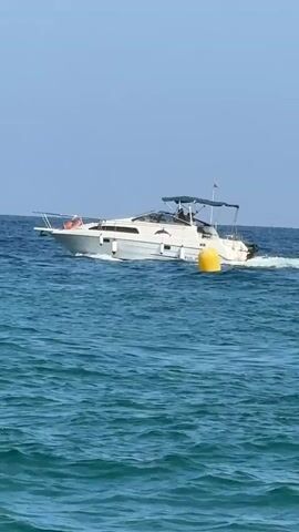 A white motorboat with a dark canopy navigates through blue ocean water under a clear sky. The boat has a cabin, a windshield, and an outboard motor. A yellow buoy is visible near the stern of the boat. In the foreground, the ocean is a vibrant turquoise with visible waves. The scene transitions to a sandy beach where two people are sitting. They are facing the water, with one person wearing dark swim trunks and the other wearing a light-colored swimsuit. The water at the edge of the beach is shallow and clear. Another person, partially submerged in the water, appears to be swimming or snorkeling. In the distance, another boat is visible on the horizon.