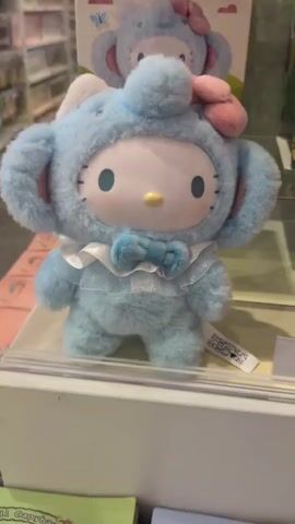 The video shows a close-up of a light blue plush toy resembling Hello Kitty dressed in an elephant costume. The toy has a white face with black eyes and a yellow nose. It is wearing a blue bow tie and a frilly white collar. A pink bow is on top of its head. The toy is displayed on a white shelf or counter, with a blurry background of shelves stocked with items. A person's hand, with fingers extended, enters the frame from the right and gently touches the toy's right side. The hand remains in the frame for the duration of the short video, with the fingers subtly moving.