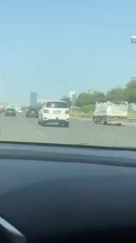 The view from inside a car driving on a highway is shown. The sky is bright and clear. Several other cars are on the road including a white SUV that is in the lane ahead. Some light industrial structures and some trees are seen on the right-hand side of the road. Some tall buildings are seen in the distance.