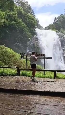 The video captures a wide shot of a tall, multi-tiered waterfall cascading down a rocky cliff face. Lush green trees and vegetation surround the waterfall, and the sky above is a mixture of blue with scattered white clouds. In the foreground, a wooden boardwalk with some water on its surface leads the viewer towards the waterfall. A single person, seen from behind, stands on the boardwalk, facing the waterfall. They are wearing a light-colored t-shirt, dark shorts, and light-colored sneakers. The person appears to be holding a phone or camera and is either taking a picture or recording the scenic view. A dark wooden railing separates the boardwalk from a grassy area in front of the waterfall. There are also some signs or posts visible behind the person. The overall scene is one of natural beauty and a person appreciating the environment.
