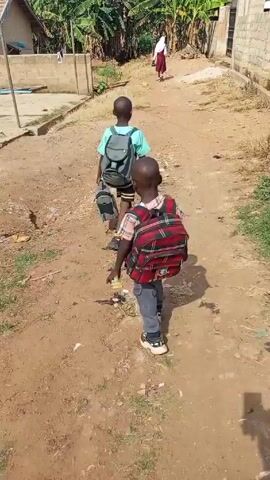 Two young boys walk along a dirt road. They are both wearing backpacks and carrying bags. One boy has a red plaid backpack and the other has a light blue backpack. A girl in a white top and maroon skirt walks ahead of them.  The road is lined with buildings and vegetation. The area appears to be rural.