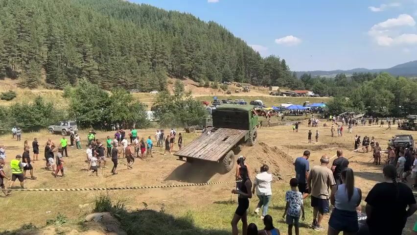 A large crowd of people watches as a truck drives up a dirt ramp. The truck is green and appears to be old, with a flatbed trailer attached. The people are dressed in casual clothing, and many are holding up their phones to take pictures or videos. The event appears to be taking place outdoors, with a wooded hill in the background. The weather is sunny and clear. There are several other vehicles parked in the background, including what appear to be other trucks and SUVs. There are also tents set up, suggesting that this might be some kind of event or festival.