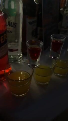 The video shows a close-up, slightly blurred view of a bar counter with several shot glasses filled with brightly colored liquids. In the foreground, there are multiple glasses filled with a yellow liquid, likely a juice or liqueur. Stacked on top of some of these yellow-filled glasses are shot glasses containing a red liquid. Further back, more shot glasses are visible, some with red liquid and others with a blue liquid. Behind the glasses, bottles of alcohol are lined up on the counter, including what appears to be gin, and other spirits. The lighting suggests a dimly lit bar environment. No people are clearly visible in the frame. The focus is on the arrangement of drinks, suggesting a preparation or presentation of shots.