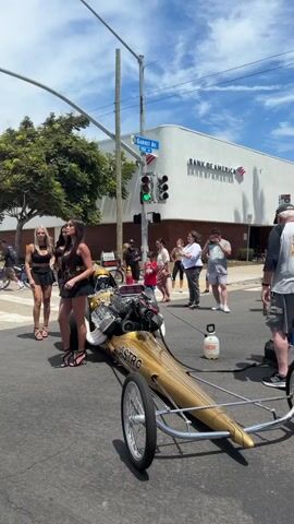 The video captures a street scene on a sunny day, likely a parade or event. In the foreground, a gold dragster with a visible engine and the word "ASTRO" on its side is positioned in the middle of the street. Three women in black outfits and high heels stand to the left of the dragster, appearing to pose or be part of the display. Behind them, several other people are gathered, some observing the dragster and the women. A traffic light is visible, showing a green signal. In the background, a Bank of America building with a distinct white, curved facade stands prominently. A street sign for "Garnet Ave" is also visible. Further in the background, more onlookers are present, and a tree provides shade. The overall atmosphere suggests a public gathering or promotional event.