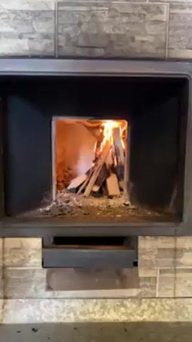 The video shows a close-up view of a fireplace with a fire burning inside. The fireplace is framed by decorative, gray and white square tiles, and it has a black metal interior. Inside, several pieces of wood are stacked vertically, engulfed in flames. Ash and small debris are scattered on the bottom of the fireplace. The camera zooms slightly inward to get a closer view of the fire.
