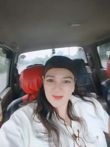 The video shows a woman with long dark hair and a bandana covering her hair. She is wearing a white jacket and red lipstick. She is sitting in the passenger seat of a car, looking at the camera and making facial expressions. The interior of the car is visible, including the red and black seats behind her. The scene appears to be during the day, with some light coming through the windows. The woman's head moves slightly throughout the clip.