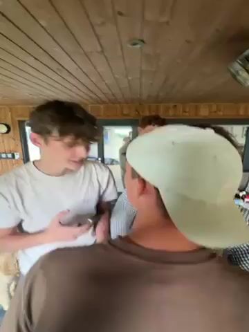 Four young men are gathered inside a room that looks like a sunroom or enclosed porch. The room has wood paneling on the ceiling and walls, with large windows along one side. In the middle of the room, there is a large picnic-style table covered with an assortment of bottles and containers. One of the men, wearing a white t-shirt and light blue pants, is holding a phone and talking. He picks up a green bottle from the table. Another man, with red hair, stands in the background, and a third man, wearing a striped shirt, is partially visible. The fourth man, closest to the camera, wears a light brown t-shirt and a white baseball cap. A light-colored couch is visible in the background.